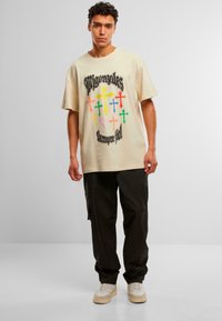 Camiseta de gran tamaño beige con gráficos de cruces coloridos y texto negro, combinada con pantalones cargo oscuros y zapatillas beige.