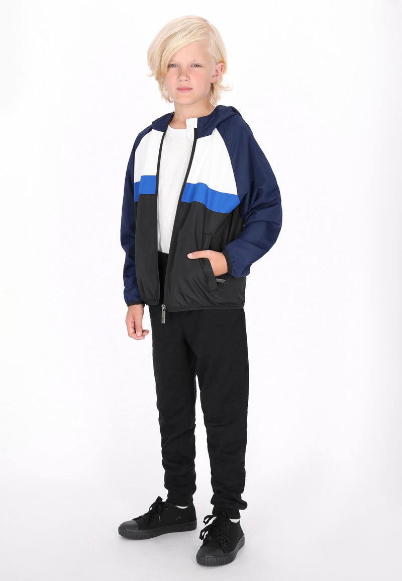 Schwarze und marineblaue Windjacke mit horizontalen weißen und blauen Streifen, Reißverschluss vorne und Kapuze; kombiniert mit schwarzen Hosen und schwarzen Turnschuhen.