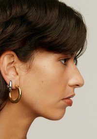 Pendientes de aro en oro y plata con un acabado brillante; el diseño presenta un aro grueso y un acento más pequeño conectado.