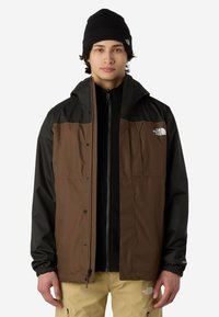 Brązowa i czarna wodoodporna kurtka z kieszenią z przodu, zamkiem błyskawicznym i elastycznymi mankietami; noszona na czarnej kurtce na zamek; w zestawieniu z beżowymi spodniami.