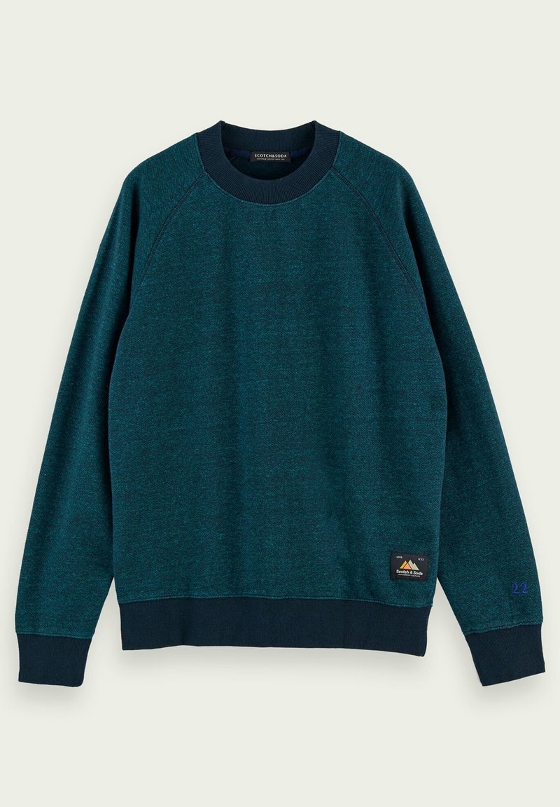 Sudadera en verde azulado oscuro con mangas raglán, cuello y puños de canalé, presenta un logotipo de parche en la parte inferior izquierda y detalles en púrpura en la manga.