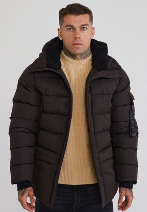 Homme avec des tatouages et un anneau au nez portant une doudoune noire par-dessus un pull en maille beige, debout devant un fond gris uni.