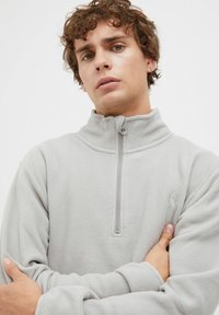 Heller grauer Fleece-Pullover mit hohem Kragen und halb-langer Reißverschluss. Verfügt über eine glatte Textur und ein kleines Logo auf der Brust.