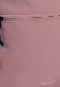 Sac à dos en tissu rose avec une surface texturée, comportant une fermeture zippée en haut, un cordon noir, et un logo de marque dans le coin inférieur droit.