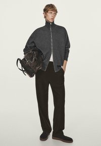Felpa grigia con zip e colletto alto, borsa marrone, pantaloni neri larghi e scarpe scure. Design semplice e casual con hardware minimo.
