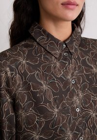 Brązowa koszula w kwiatowy wzór z kołnierzykiem, zapinana na guziki z przodu, wykonana z teksturowanej tkaniny z kontrastującymi kremowymi konturami kwiatów.