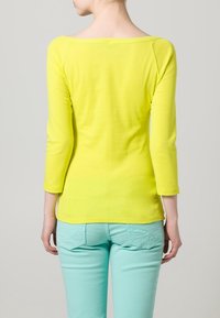 Světlá žlutá halenka s dlouhým rukávem a širokým výstřihem, doplněná světle mátovými kalhotami s kapsami na zadní straně. Hladká textura, volný střih.