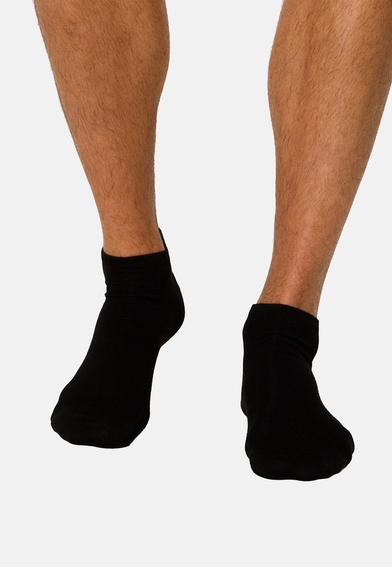 Beine mit schwarzen Knöchelsocken, die vor einem schlichten weißen Hintergrund stehen und die Passform sowie die Textur der Socken zeigen.