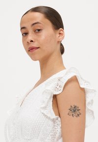 Hvit blondekjole med volangerm, med et teksturert stoff og dyp V-utringning. En blomstertatovering er synlig på overarmen.