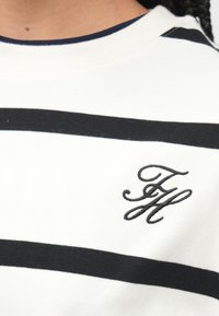 Weißer Pullover mit breiten schwarzen horizontalen Streifen, vorne ein gebogenes "FH"-Logo in schwarzer Stickerei. Weiche Baumwollstruktur.