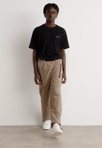Black short-sleeve t-shirt with logo, paired with beige cargo pants featuring side zippers and white sneakers. Plain background.