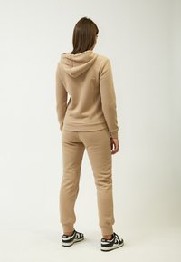 Beige Hoodie und Jogginghose aus weichem Stoff, mit einer Kapuze mit Kordelzug und gerippten Bündchen, kombiniert mit schwarzen und weißen Sneakern.