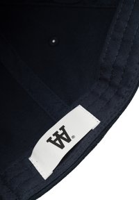 Sötétkék szövet sapka belseje, fehér címkével, amelyen fekete logó található. A széleken varrás található, valamint egy kis gomb.