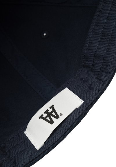 Interno di un cappello in tessuto blu scuro con un'etichetta bianca che mostra un logo nero. Presenta cuciture lungo i bordi e un piccolo bottone.