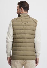 Beige gesteppte Weste aus leichtem Nylonmaterial, mit hohem Kragen und gerippten Strickärmeln. Der Rücken verfügt über ein dreieckiges Yoke-Detail.