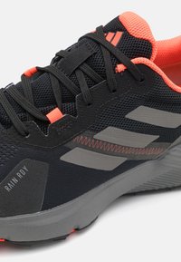 Scarpe da ginnastica nere con accenti grigi, tessuto in rete e dettagli arancioni. Presentano una suola arrotondata e un tallone rinforzato per il supporto.
