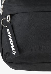 Černá látková taška s texturovaným povrchem, která má stříbrný zip a obdélníkovou klíčenkovou cedulku s nápisem "CONVERSE" v bílé barvě.