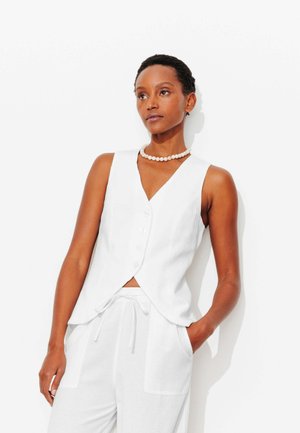 Femme aux cheveux courts portant un gilet blanc sans manches boutonné, un pantalon blanc avec cordon de serrage, un collier de perles, debout avec une main dans la poche.