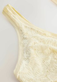 Kremfarvet blonde-g-streng med blomsterdesign og en blød elastisk taljebånd på en ensfarvet baggrund.