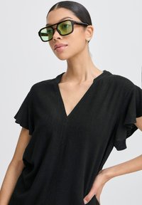 Top nero con scollatura a V, maniche corte a volant e tessuto testurizzato. Il modello indossa occhiali da sole oversize con tonalità verde, che evidenziano un design elegante.