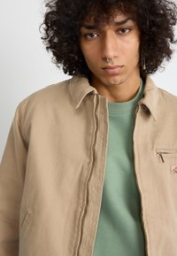 Tan canvas jacket with a collared neck and zip closure, layered over a green sweatshirt. Features a small pocket with a zipper.