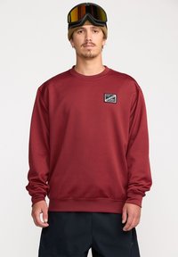 Sweatshirt bordeaux à manches longues, avec des poignets et un ourlet côtelés, et un petit logo en patch noir en haut à gauche. Porté avec des lunettes de ski.