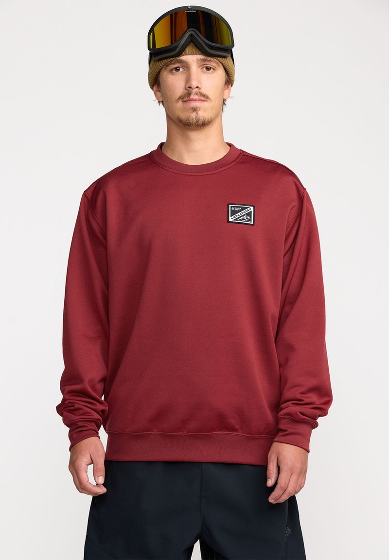 Sweatshirt bordeaux à manches longues, avec des poignets et un ourlet côtelés, et un petit logo en patch noir en haut à gauche. Porté avec des lunettes de ski.