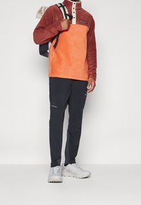 Sudadera de forro polar Columbia en naranja y óxido, con un cuello alto y cierre de botones. Combinada con pantalones oscuros que tienen cremalleras y acentos blancos.