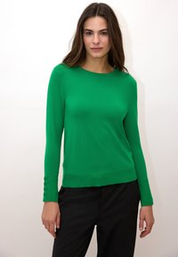 Pull vert à manches longues en tissu doux, avec un col rond et des boutons sur les poignets, associé à un pantalon foncé.