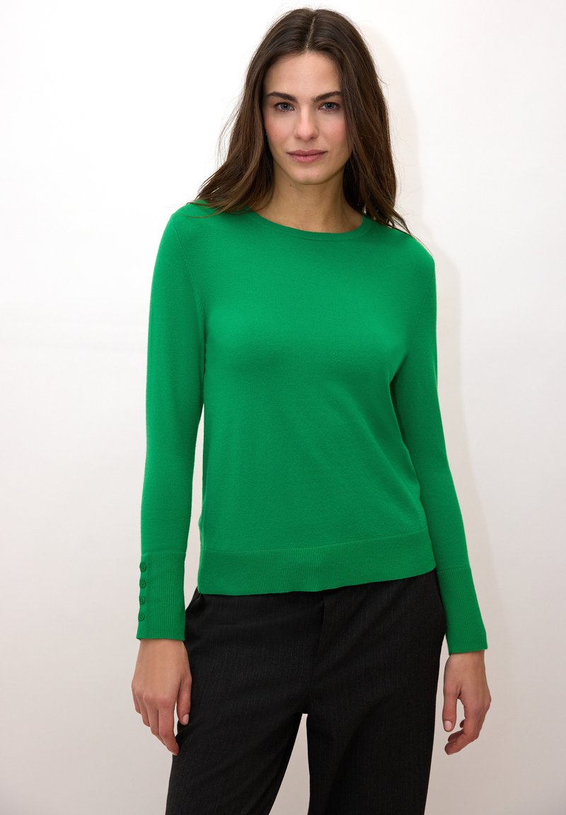 Pull vert à manches longues en tissu doux, avec un col rond et des boutons sur les poignets, associé à un pantalon foncé.