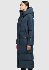 Abrigo largo de plumas azul marino con capucha grande, diseño acolchado, bolsillos laterales con cremallera y silueta ajustada. Acabado texturizado con un branding mínimo.