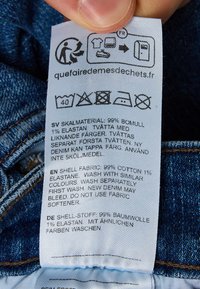 Etykieta pielęgnacyjna odzieży przedstawiająca skład tkaniny, symbole prania oraz instrukcje w języku szwedzkim, angielskim i niemieckim na materiale dżinsowym.