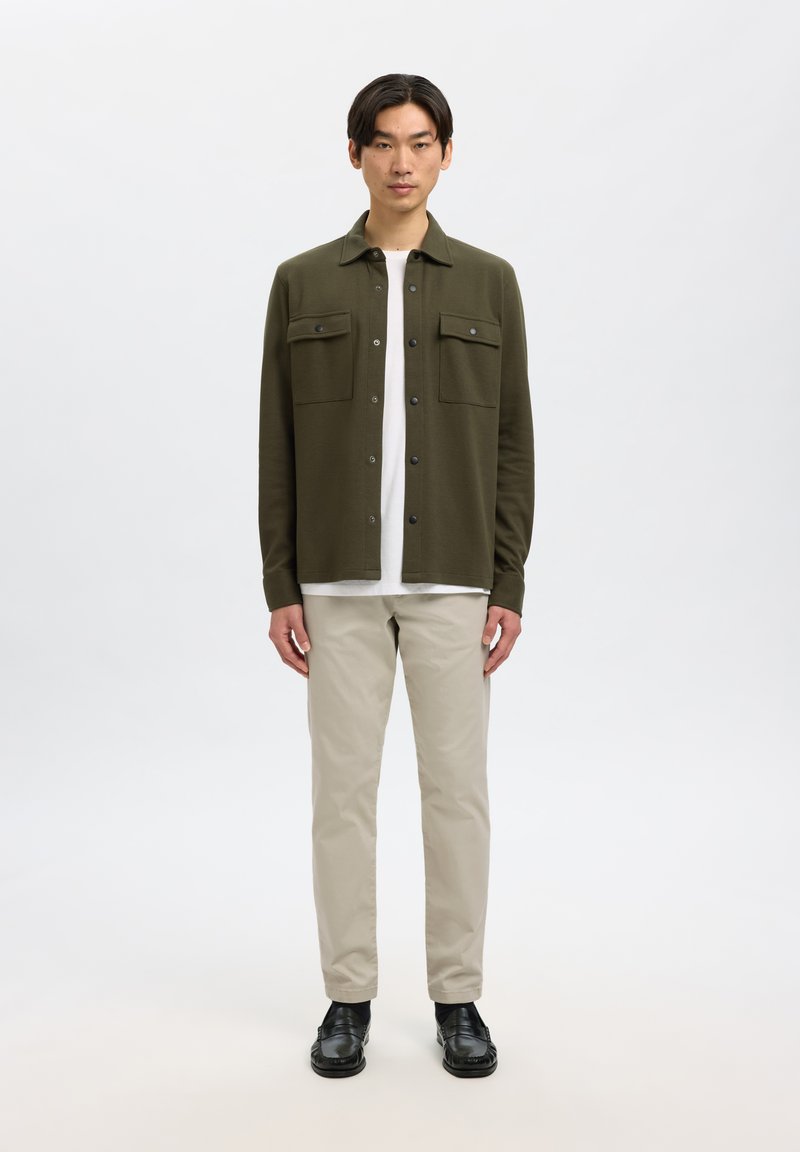 Man staat, draagt een olijfgroen overhemdjasje met knopen, wit ondershirt, beige broek en zwarte schoenen tegen een effen lichte achtergrond.
