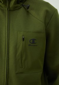 Sweat à zip couleur olive fabriqué en tissu doux, avec une poche poitrine zippée et un logo Champion noir sur le devant.