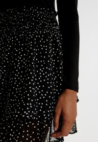 Falda negra con lunares, de tejido texturizado, con cinturilla elástica fruncida, diseño en capas y patrones circulares blancos. Mano visible a un lado.