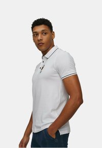 Polo shirt bianca con un design semplice, rifiniture nere lungo il colletto e le maniche, e un piccolo logo sul petto. Texture liscia.