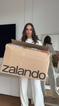 Smeđi kartonski kutija s Zalando logotipom, djelomično otvorena, drži je osoba u bijelom odijelu. U pozadini se nalaze ogledalo i namještaj.