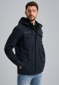 Giacca a vento navy con cappuccio, dotata di molteplici tasche frontali, chiusura zip e polsini a costine; tessuto liscio, decorata con toppe sulle spalle.
