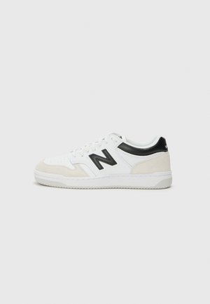 Hvid og beige lavtop-sneakers med sort "N" logo og sort hælkappe, snørestil og flad gummisål på en lysegrå baggrund.
