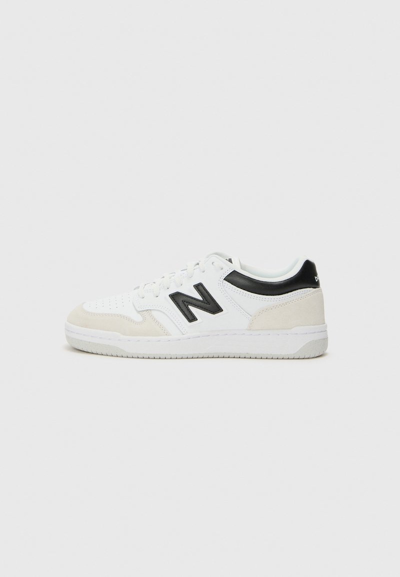 Baskets basses blanches et beige avec logo "N" noir et languette noire au talon, style à lacets et semelle en caoutchouc plate sur fond gris clair.