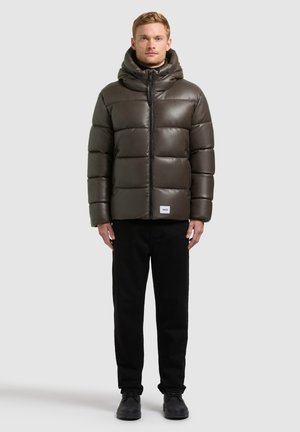 Uomo che indossa una giacca imbottita verde oliva con cappuccio, pantaloni neri e scarpe nere in piedi davanti a uno sfondo chiaro e uniforme.