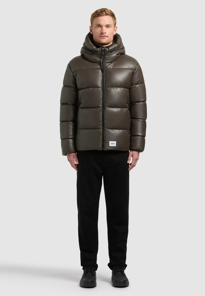 Homme portant une veste matelassée kaki avec capuche, pantalon noir et chaussures noires, debout devant un fond clair uni.