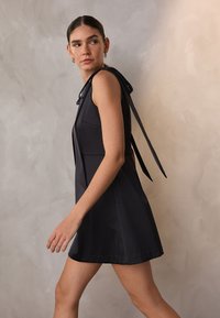 Musta hihaton mekko, jossa on tiukka vartalonmyötäinen yläosa, leveä helma ja nauhakiinnitys takana. Sileä kangas, jossa on hienovarainen kiilto.