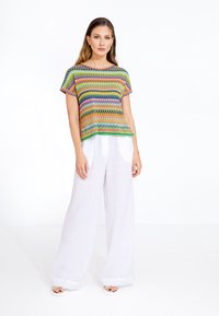 Top a maglia multicolore con motivi geometrici; maniche corte; indossato con pantaloni in lino bianchi a gamba larga; modello in piedi su uno sfondo bianco.