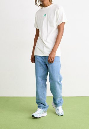 Biała bawełniana koszulka z zielonym logo Nike, niebieskie luźne dżinsy i białe trampki z akcentami w kolorze turkusowym, stojące na zielonej podłodze.