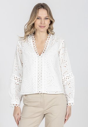 Kobieta ubrana w białą bluzkę z długim rękawem z ażurowym wzorem i falowanymi brzegami oraz beżowe spodnie, stojąca na tle jasnego, jednolitego tła.