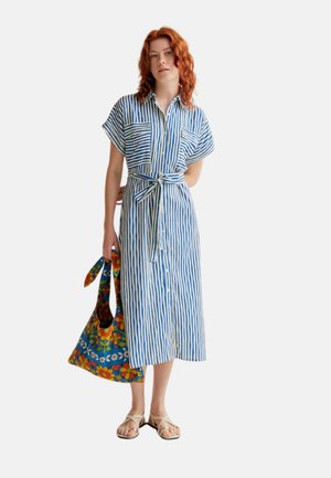 Mujer con cabello rojo que lleva un vestido midi de rayas verticales azules y blancas con cinturón, sostiene un bolso tote floral colorido y lleva sandalias beige.