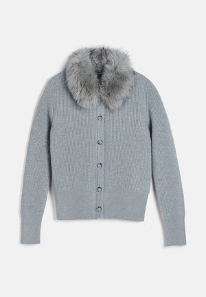 Femme portant un cardigan gris boutonné avec col en fourrure, top blanc, ceinture noire, jean large sombre et chaussures noires, debout devant un fond blanc.