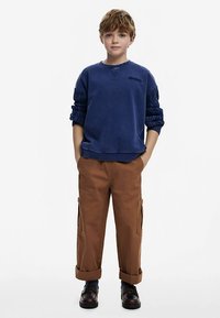 Tmavomodrá mikina s textúrovanými nafúknutými rukávmi, doplnená o hnedé cargo nohavice vyhrnuté na konci. Model stojí s rukami vreckách a má na sebe čierne topánky.