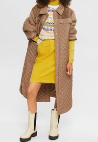 Manteau long matelassé marron avec un col, un pull coloré à motifs en dessous, une jupe jaune et des bottines beiges avec des panneaux latéraux noirs.